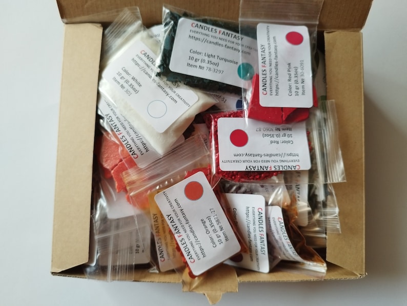 May include: A box filled with small bags of candle wax dye and fragrance. The bags are labeled with the brand name "Candles Fantasy" and the color or scent of the product. The colors include red, orange, white, turquoise, and black.