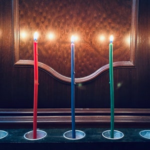 May include: Five lit candles in a row, each in a different color: yellow, red, blue, green, and black. The candles are in individual holders on a dark wooden surface.