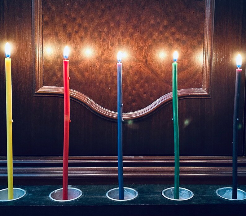 May include: Five lit candles in a row, each in a different color: yellow, red, blue, green, and black. The candles are in individual holders on a dark wooden surface.
