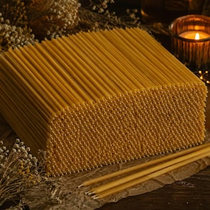 May include: A large, rectangular block of thin, golden-yellow beeswax taper candles arranged in a dense, vertical grid. Several individual matching taper candles lie horizontally in the foreground on a piece of brown paper.