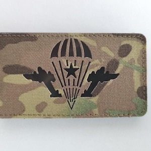 May include: A rectangular patch with a camouflage pattern. The patch features a black parachute design with a star and two stylized wings. The edges are stitched with a brown thread.