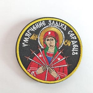 Peut inclure: Écusson brodé représentant une icône religieuse avec un halo, portant un voile rouge et tenant des flèches. Le patch comporte du texte en cercle autour de l'image, sur fond noir avec une bordure jaune.