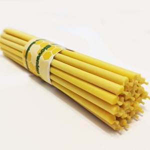 May include: A bundle of slender, yellow beeswax candles. The candles are tied together with a band featuring a honeycomb design and the text "Slovenian". The candles have a slightly textured appearance and are arranged in a neat bundle.