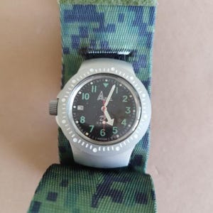 Puede incluir: Un reloj de pulsera plateado con esfera negra y números blancos. El reloj tiene una pantalla de fecha y está sujeto a una correa de camuflaje verde y azul. Las manecillas del reloj son blancas.