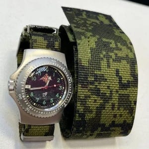 Puede incluir: Un reloj de pulsera plateado con esfera negra, números verdes y un emblema de estrella roja. Tiene una correa de camuflaje verde y negro. Se muestra una correa de camuflaje a juego junto al reloj.