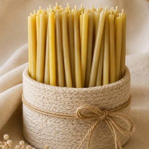 Puede incluir: Una colección de velas de cera de abeja amarillas, finas y cilíndricas, colocadas en posición vertical dentro de una cesta redonda de cuerda trenzada de color crema. La cesta está envuelta con un cordel de yute atado en un lazo.