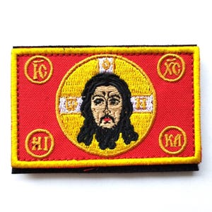 Puede incluir: Parche bordado que representa un icono religioso con halo, cabello oscuro y barba, sobre un círculo amarillo. El parche está sobre un fondo rectangular rojo con ribete amarillo y texto religioso en cada esquina.