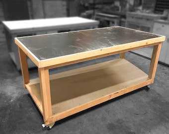 Puede incluir: Un banco de trabajo de madera con una tapa de metal y un estante inferior. El banco de trabajo está sobre ruedas y tiene un acabado de madera clara.