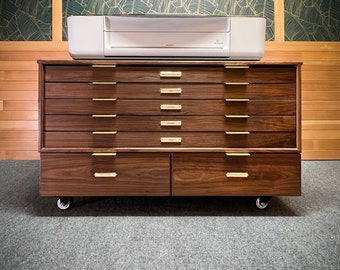 Op de afbeelding: Een donkerhouten kast met acht laden en messing handgrepen. De kast heeft wielen en is bedekt met een witte printer.