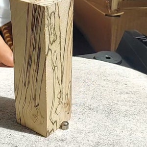 May include: A rectangular wooden block with a light beige base colour and dark brown grain patterns. A small, silver-coloured metal object rests near the base of the block. The wood has a natural, organic appearance.