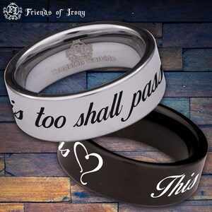 May include: Two tungsten carbide rings. The top ring is silver with black lettering that reads "This too shall pass". The bottom ring is black with a white heart and white lettering that reads "This".