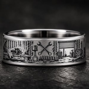 May include: A silver-toned metal ring featuring a detailed laser-engraved wrap-around design of an automotive garage scene. The engraving depicts various tools, a workbench, a piston, crossed wrenches, and a vehicle tire within a workshop.
