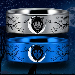 May include: Two tungsten carbide rings featuring a wolf's head and crescent moon design. The top ring is silver with a black wolf and moon. The bottom ring is blue with a black wolf and moon.