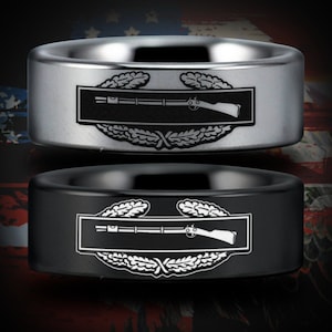 May include: Two tungsten carbide rings with a black and silver finish. The rings feature a military insignia with a rifle and laurel wreath design.