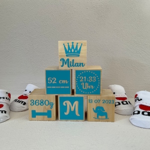 Peut inclure: Des blocs en bois avec des inscriptions bleues et dorées indiquant "Milan", "52 cm", "21:33 Uhr", "3680g", "M" et "13 07 2023". Les blocs sont empilés en forme de pyramide. Il y a deux chaussettes de bébé blanches avec des cœurs rouges.