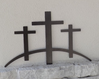 May include: Three black metal crosses are mounted on a curved metal base, creating a silhouette of a hill. The crosses are of different sizes, with the largest in the center.