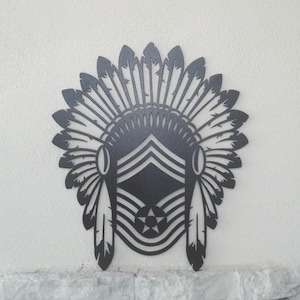 May include: A dark gray metal wall art piece in the shape of a Native American headdress. The headdress features a series of feathers, a central emblem with stripes and a star, and two hanging feathers on the sides. The art is mounted on a white wall.