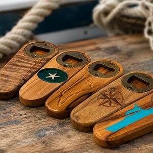 Könnte beinhalten: Fünf Flaschenöffner aus Holz, die in einer Reihe angeordnet sind. Jeder hat eine abgerundete rechteckige Form mit einem messingfarbenen Metallöffner, der mit zwei Schrauben befestigt ist. Die Designs umfassen Holzmaserung, eine grüne Einlage mit Stern, eine Landkarte, einen Kompass und ein Boot.