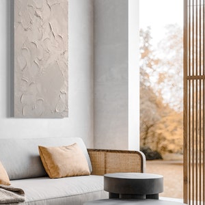 Puede incluir: Una pintura de lienzo texturizado gris claro cuelga de una pared blanca sobre un sof&aacute; gris claro con un reposabrazos de mimbre marr&oacute;n. El sof&aacute; tiene dos almohadas, una blanca y una naranja. Dos mesas nido redondas grises est&aacute;n delante del sof&aacute;. El fondo es una vista borrosa de &aacute;rboles a trav&eacute;s de una ventana.