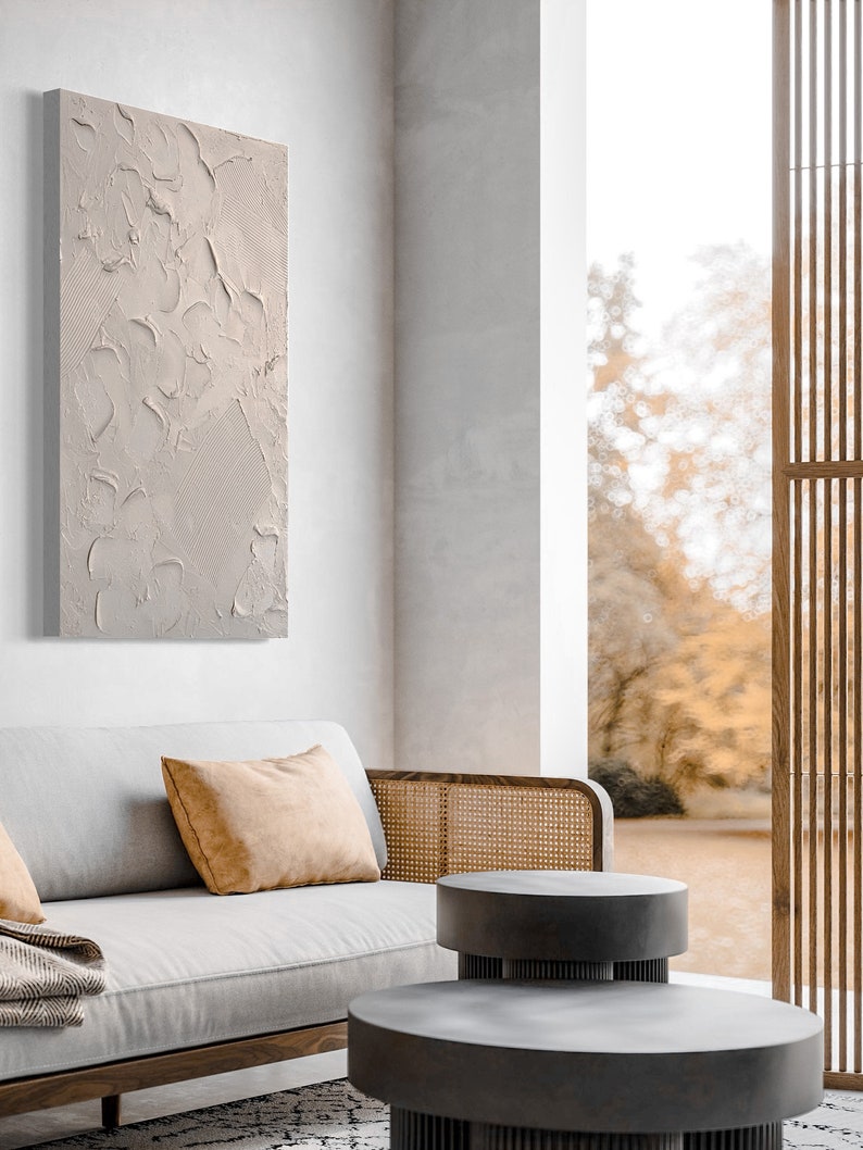Puede incluir: Una pintura de lienzo texturizado gris claro cuelga de una pared blanca sobre un sof&aacute; gris claro con un reposabrazos de mimbre marr&oacute;n. El sof&aacute; tiene dos almohadas, una blanca y una naranja. Dos mesas nido redondas grises est&aacute;n delante del sof&aacute;. El fondo es una vista borrosa de &aacute;rboles a trav&eacute;s de una ventana.