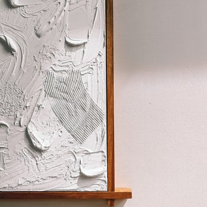 Puede incluir: Una pintura abstracta blanca con pinceladas texturizadas, enmarcada en un marco de madera natural.