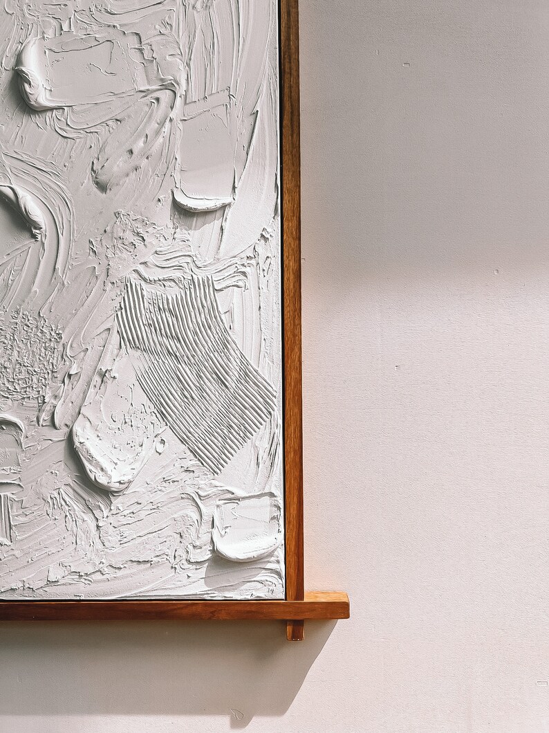 Puede incluir: Una pintura abstracta blanca con pinceladas texturizadas, enmarcada en un marco de madera natural.
