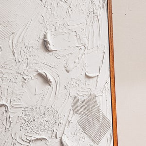 Puede incluir: Una pintura abstracta texturizada blanca en un marco de madera. La pintura tiene una textura en relieve, tridimensional.