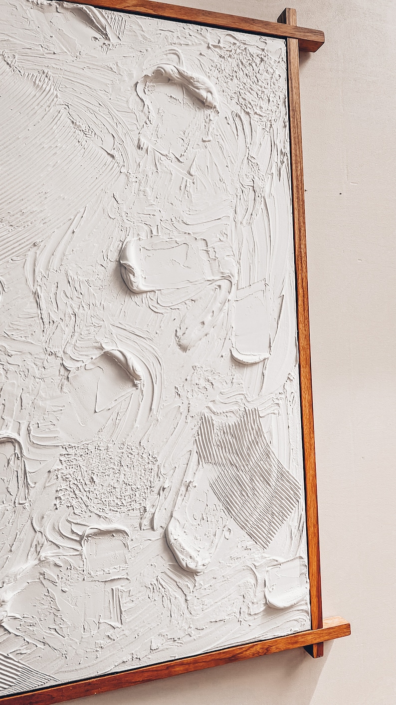 Puede incluir: Una pintura abstracta texturizada blanca en un marco de madera. La pintura tiene una textura en relieve, tridimensional.