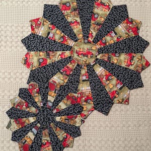 May include: Two quilted, starburst-shaped placemats. The placemats feature a repeating pattern of black and white circles, and a fabric with red trucks, pumpkins, and fall foliage. The placemats are arranged on a cream-colored, textured surface.