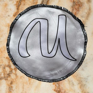 Può includere: Un cerchio di raso bianco con un bordo cucito nero. Il cerchio presenta una lettera "M" bianca in un carattere stilizzato.