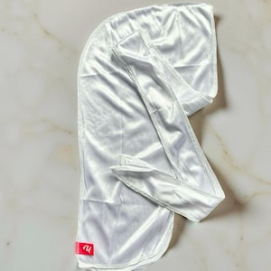 May include: A white satin durag, a head covering with a long tail and flaps. The durag is made of a shiny, smooth fabric. A small red tag with a logo is visible on the bottom.