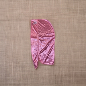 May include: A pink satin durag with a shiny, reflective surface. The durag has a curved top and long, flowing tails. A small, silver-colored emblem is visible on the front of the durag.
