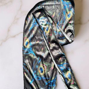 May include: Black satin durag with a vibrant peacock feather pattern in shades of blue, green, yellow, and pink. The durag features a white stitched edge and a white logo on the front.