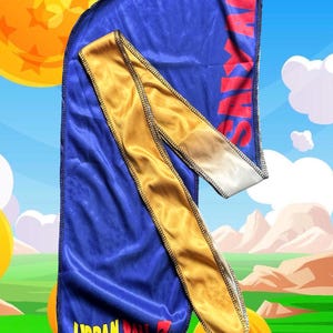 Könnte beinhalten: Ein blauer Durag mit goldener Zierleiste und einem goldenen Kopftuch. Der Durag trägt den Text "URBAN BALL Z VEGETA SAGA" in Gelb und Rot. Das Wort "SAYIAN" ist rot auf der Oberseite des Durags aufgedruckt.