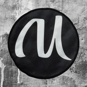 Può includere: Un cerotto circolare bianco e nero con una lettera "M" stilizzata bianca al centro.