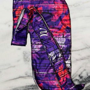 Puede incluir: Un pañuelo de cabeza de satén morado y rosa con un patrón de pared de ladrillo y un diseño de graffiti. El pañuelo tiene un ribete de satén blanco y la letra "M" está bordada en el lateral.