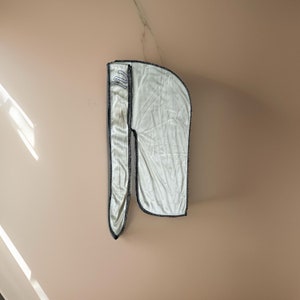 Può includere: Un durag di raso bianco con un bordo nero. Il durag ha un piccolo disegno ricamato sul davanti.