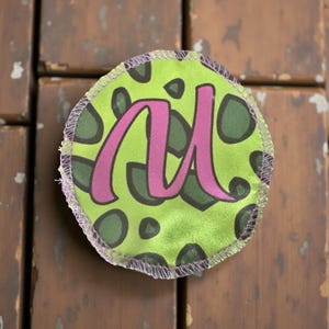 Può includere: Una toppa rotonda in tessuto con sfondo verde lime e pois verde scuro. Una grande lettera "M" magenta in corsivo è centrata sulla toppa. La toppa ha un bordo cucito lilla chiaro.