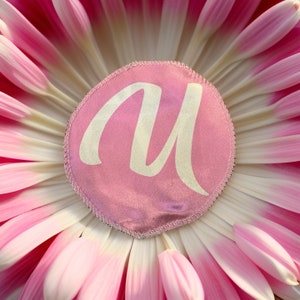 Può includere: Un cerchio di tessuto rosa con una lettera "M" bianca al centro, circondato da petali di fiori rosa.