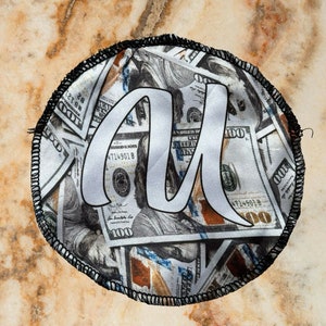 Può includere: Una toppa rotonda con un bordo nero e uno sfondo bianco. La toppa è ricoperta da immagini di banconote in dollari americani. La lettera "A" è scritta in bianco sopra le banconote in dollari.