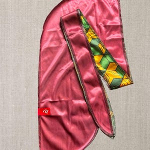 May include: A burgundy satin durag with a patterned accent. The accent features a geometric design in yellow, green, and black. A small red tag with white text is attached to the durag.