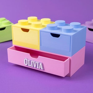 Può includere: Un organizzatore a cassetti multicolore in rosa, giallo, blu e verde, a forma di blocchi Lego. Il cassetto inferiore è aperto e ha il nome "Olivia" scritto su di esso.