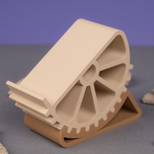 Puede incluir: Un soporte para teléfono beige con un diseño de engranaje. El soporte está hecho de plástico y tiene una pequeña plataforma para sostener un teléfono. El soporte está inclinado para sostener el teléfono en un ángulo de visión cómodo.