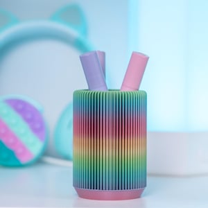 May include: A rainbow-colored cylindrical container with three pastel-colored lip balms inside. The container is made of plastic and has a ribbed texture. The lip balms are arranged vertically in the container.