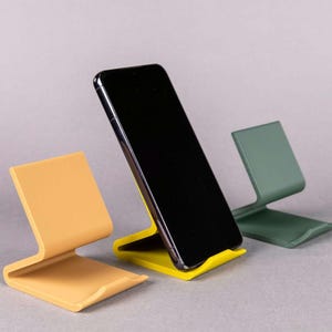 Op de afbeelding: Drie telefoonstandaards in verschillende kleuren: geel, oranje en groen. De standaards zijn gemaakt van plastic en hebben een eenvoudig ontwerp. Een zwarte smartphone ligt op de gele standaard.