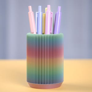 Może przedstawiać: Stojak na długopisy w kolorze tęczy z żebrowanym wzorem. Sześć długopisów w różnych pastelowych kolorach stoi pionowo w stojaku.