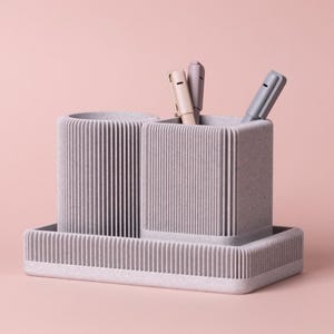 May include: A light grey desk tidy with a ribbed design, holding three pens. The organiser has two compartments and a rectangular base. The pens are beige, grey, and light grey. The background is a solid light pink.