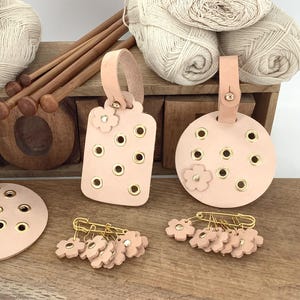 May include: A collection of craft supplies including light pink leather stitch markers, in round and rectangular shapes, with gold eyelets and flower accents. Also shown are gold safety pins with flower charms, wooden knitting needles, and yarn.