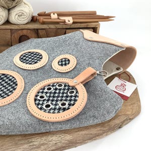 May include: A gray felt knitting bag with leather accents and houndstooth patterned inserts. The bag has a leather handle and a metal clasp. Wooden knitting needles and yarn are in the background.