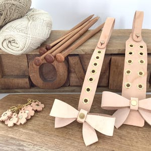 Puede incluir: Una colección de suministros para manualidades que incluye hilo, agujas de tejer de madera y accesorios de cuero. Se muestran dos correas de cuero rosa claro con detalles de lazo y ojales dorados. Una caja de madera con la palabra "LOVE" está en el fondo.